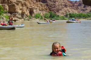 Girl swimming and smiling in the Green River with rafts and kayaks in the background in Desolation Canyon - Mild to Wild